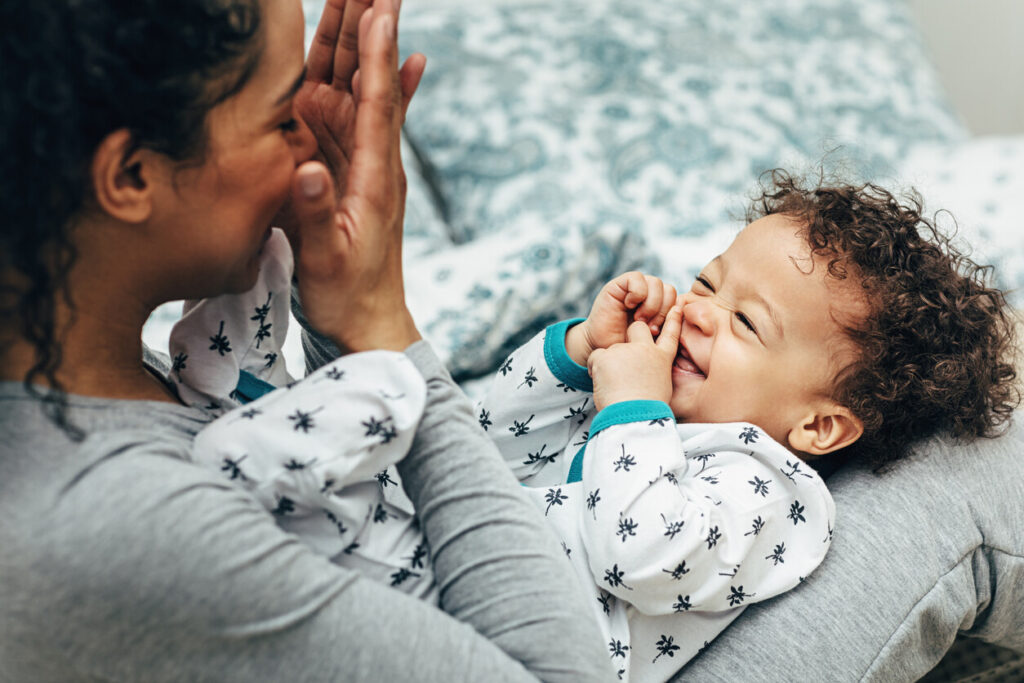 A mother playing peek-a-boo with her smiling baby boy on her lap.