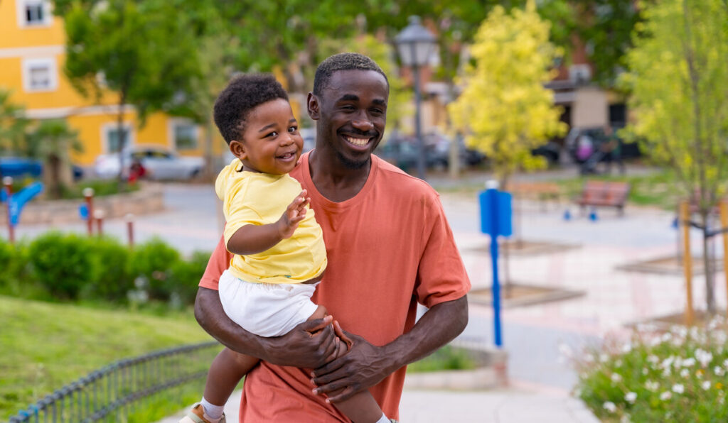 Caregiver holding young child and walking through a park, both laughing.