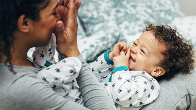 A mother playing peek-a-boo with her smiling baby boy on her lap.