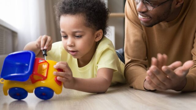 Preschool-age boy pretending to repair a toy car with a plastic wrench on the floor with his dad.