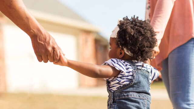 Toddler walking with two caregivers, facing away from camera