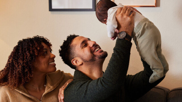 Two caregivers sitting at home, one holding infant above their head