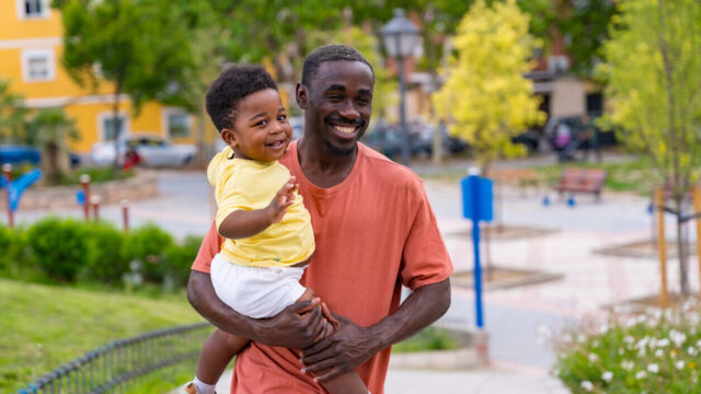 Caregiver holding young child and walking through a park, both laughing.